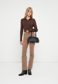 Brun ribbet langærmet polo, beige fløjl flagrende bukser, og sorte hælede ankelstøvler. Holder en lille sort håndtaske. Ensfarvet baggrund.