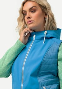 Gilet blu con cappuccio, caratterizzato da un materiale esterno liscio e dettagli imbottiti. Maniche verde testurizzato si estendono dal corpo. Dettaglio della cerniera bianca.