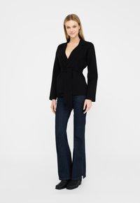 Sort indpakket blazer med dyb V-udskæring, lange ærmer og en knyttet talje. Sæt med mørke flared jeans og sorte ankelstøvler.