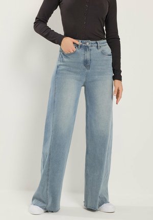 Person wearing light blue wide-leg jeans and black long-sleeve buttoned top, posing with one hand in pocket and white shoes.