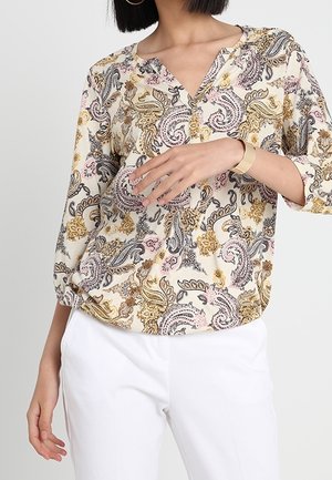 Femme portant une blouse à manches longues à motif paisley dans des tons crème, rose, or et noir, associée à un pantalon blanc et un bracelet doré.