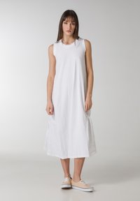 Vestido maxi blanco sin mangas con un corte holgado, que incluye bolsillos laterales y un dobladillo inferior ligeramente recogido, combinado con zapatillas de lona beige.