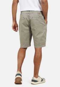 Pantaloni corti kaki da uomo, in cotone, con taglio dritto, tasche laterali e posteriori, abbinati a una camicia bianca e scarpe da ginnastica neutre.