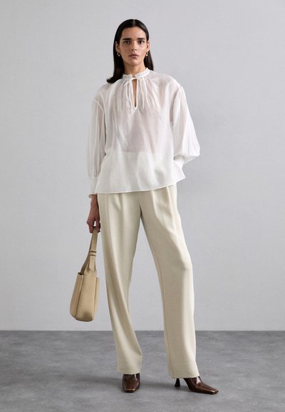 Blouse blanche, ample, avec un col en découpe et un col froncé, associée à un pantalon crème. Talons marron à bout pointu et sac beige.