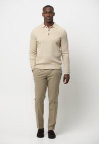 Beige gerippter Strickpullover mit einem Kragen und einer Knopfleiste mit drei Knöpfen, kombiniert mit hellbeigen Hosen und dunkelbraunen Mokassins.