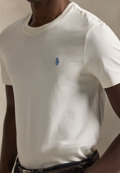 T-shirt en coton crème avec un col rond classique, présentant un petit logo polo bleu brodé sur la poitrine. Porté avec une ceinture foncée.