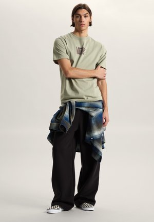 Giovane uomo in piedi con le braccia incrociate, indossa una t-shirt beige Vans, pantaloni neri larghi, scarpe slip-on a quadri e una camicia a quadri blu legata in vita.