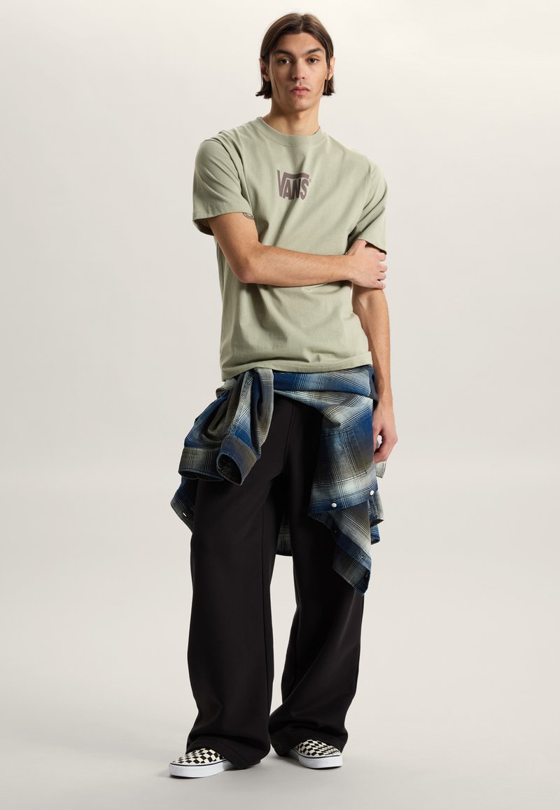 Jeune homme debout, les bras croisés, portant un t-shirt Vans beige, un pantalon large noir, des chaussures à enfiler à carreaux, et une chemise à carreaux bleue nouée.