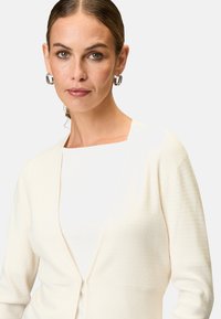 Cremefarbener Cardigan mit strukturiertem Muster, offener Vorderseite und langen Ärmeln, über einem weißen Oberteil getragen. Subtile Details betonen den Stoff.