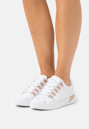 Benen met witte lage sneakers met lichtroze veterogen en subtiele multicolor geometrische patronen op een witte achtergrond.