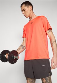 Lyseorange trænings T-shirt med rund hals og korte ærmer, parret med sorte shorts. T-shirten har ordet "HYROX" og PUMA-logoer.