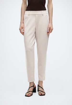 Nő, bézs színű, szabott nadrágot visel, fekete, pántos magassarkú szandállal, fehér háttér előtt állva.