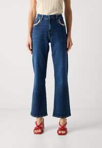 Jeans in denim a gamba larga in lavaggio scuro con vita media, caratterizzati da cuciture decorative e due tasche frontali. Abbinati a tacchi a stiletto rossi.
