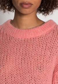 Zbliżenie na osobę w puszystym, luźno dzierganym różowym swetrze z prążkowanym okrągłym dekoltem i naturalnymi kręconymi włosami.