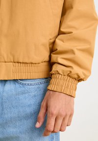 Giacca bomber beige con polsini e orlo elasticizzati, caratterizzata da una trama liscia. La mano del modello poggia su jeans in denim azzurro chiaro.