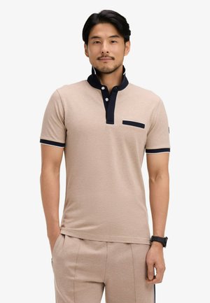 Homme portant un polo à manches courtes beige avec col noir et détail sur la poche, pantalon beige et une montre noire au poignet, debout les mains dans les poches.