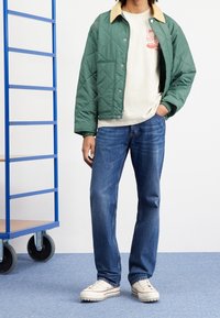 Zelená prošívaná bunda s béžovým límcem, krémová košile s grafickým potiskem, modré džíny z denimu a krémové boty s texturovanou podrážkou.