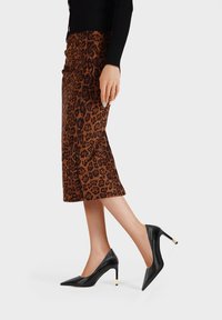 Donna che indossa una gonna midi a stampa leopardata marrone e scarpe con tacco alto nere a punta, in piedi su uno sfondo bianco.