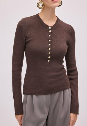 Femme portant un haut marron côtelé ajusté à manches longues avec des boutons dorés et un pantalon beige plissé, accessoirisé de grandes boucles d’oreilles créoles dorées.