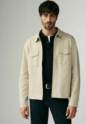 Chaqueta beige con cremallera frontal, dos bolsillos en el pecho y un diseño con cuello. Llevada sobre un polo negro y combinada con pantalones negros.