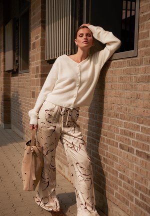 Kvinde der læner sig mod murstensvæg iført hvid knappet sweater, cremelige bukser med bladprint, holder beige taske udendørs.