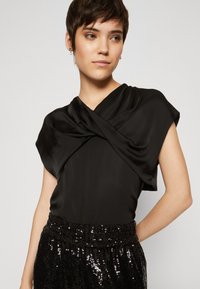 Top in satin nero con scollatura incrociata, maniche corte e silhouette aderente. Abbinato a pantaloni neri a vita alta con paillettes.