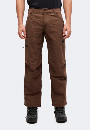 Homme portant un pantalon d’extérieur marron avec une poche cargo et des chaussures de randonnée noires, debout devant un fond clair uni.