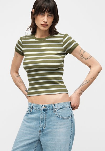 Γυναίκα με τατουάζ φοράει στενό πράσινο-άσπρο ριγέ μπλουζάκι και ανοιχτό μπλε ψηλόμεσο τζιν, στέκεται με τα χέρια στη μέση.