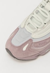 Lila Veloursleder- und Mesh-Sneaker mit einer strukturierten hellgrauen Oberseite, markanter Schnürung und dezentem Branding an der Seite. Geschwungene Designelemente.