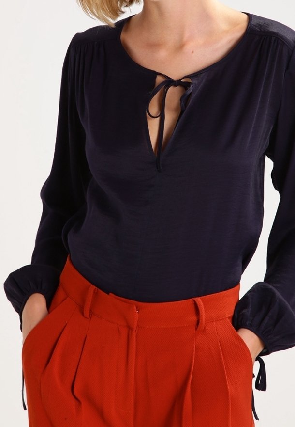 Blouse marine avec un décolleté en keyhole et un nœud, associée à un pantalon orange texturé taille haute avec des plis avant et des poches latérales.