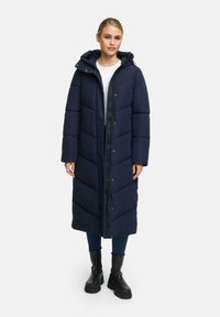 Marineblauwe lange pufferjas met chevronpatroon, rits aan de voorkant, drukknoopsluitingen en capuchon. Draag het over een witte top en in combinatie met zwarte laarzen.