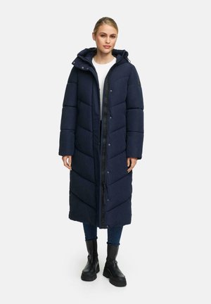 Marineblauwe lange pufferjas met chevronpatroon, rits aan de voorkant, drukknoopsluitingen en capuchon. Draag het over een witte top en in combinatie met zwarte laarzen.