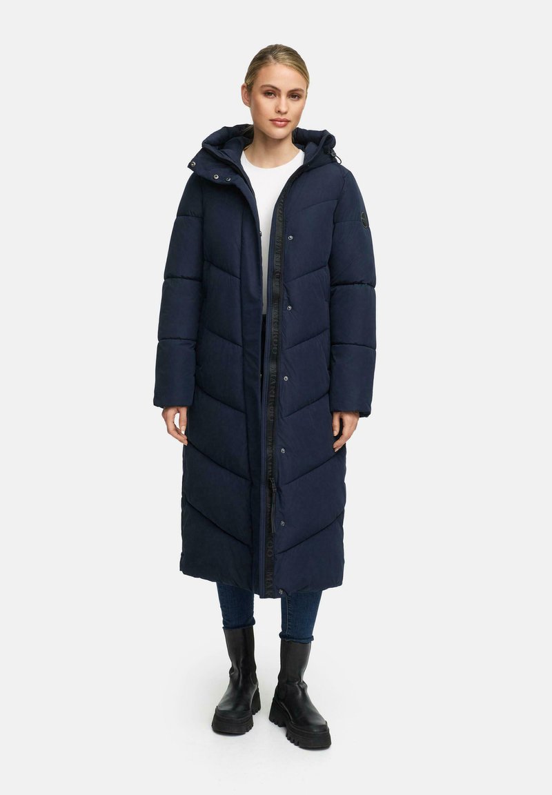 Marineblauwe lange pufferjas met chevronpatroon, rits aan de voorkant, drukknoopsluitingen en capuchon. Draag het over een witte top en in combinatie met zwarte laarzen.