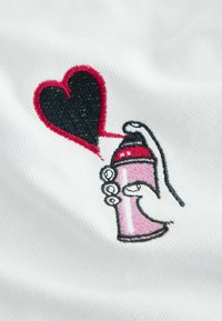 Handbesticktes Motiv einer Hand, die eine rosa Sprühdose hält und damit ein schwarzes Herz, rot umrandet, auf weißen Stoff sprüht.