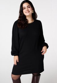 Zwarte jurk met structuur, lange pofmouwen, ronde halslijn en geribbelde stof. Draagt hierbij knielange bordeauxrode laarzen en gelaagde gouden kettingen.