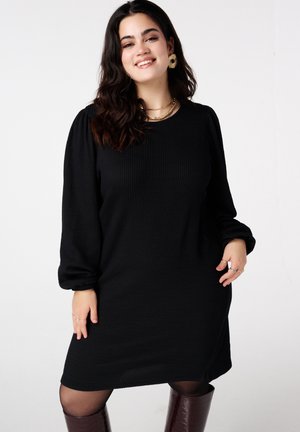 Zwarte jurk met structuur, lange pofmouwen, ronde halslijn en geribbelde stof. Draagt hierbij knielange bordeauxrode laarzen en gelaagde gouden kettingen.