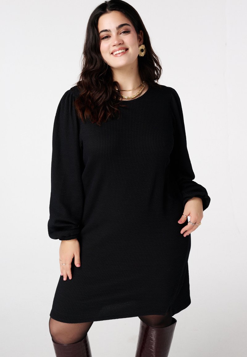 Zwarte jurk met structuur, lange pofmouwen, ronde halslijn en geribbelde stof. Draagt hierbij knielange bordeauxrode laarzen en gelaagde gouden kettingen.