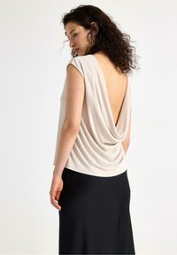 Lys beige drapering bluse med dyb V-udskæring i ryggen, korte ærmer og en blød tekstur. Kombineret med en ensfarvet sort nederdel, der fremhæver det afslappede design.