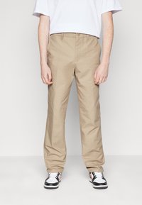 Beige cotton trousers with a straight fit, featuring a button closure, belt loops, and two side pockets. Worn with black and white sneakers.