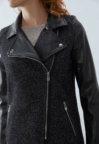 Černá a šedá bunda s rybí kostí vzorem, koženými rukávy, širokým límcem a dvojitými zipy na předních a bočních kapsách.
