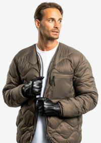 Braune, gepolsterte Steppjacke mit Reißverschluss vorne, Seitentasche und schwarzen Lederhandschuhen. Getragen über einem weißen T-Shirt.