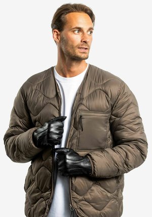 Padded brown quilted jacket with a front zip, side pocket, and black leather gloves. Worn over a white t-shirt.