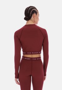 Bordó sport felső hosszú ujjakkal, rövidített hosszban, logós szegéllyel. Testhez simuló kialakítás sima textúrával és minimális varrással.