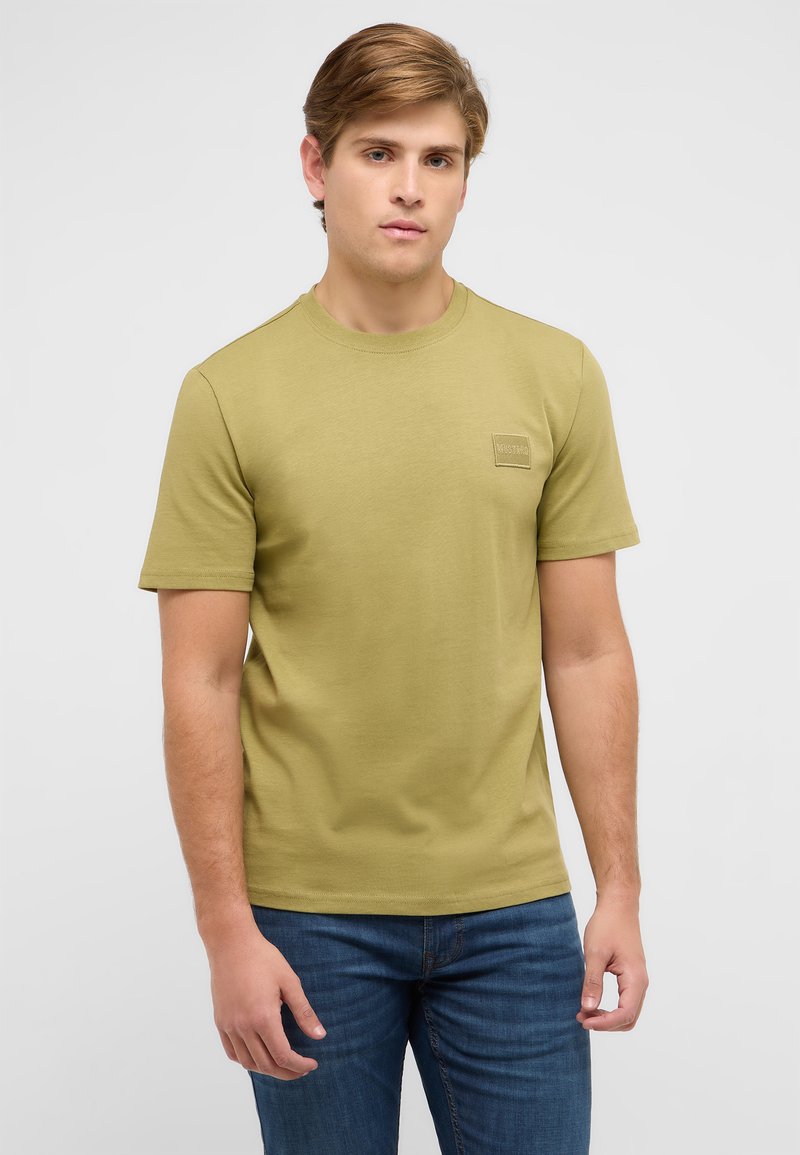 Młody mężczyzna z jasnobrązowymi włosami w oliwkowozielonym krótkim rękawie T-shircie i niebieskich dżinsach, stojący na tle jednolitego jasnoszarego tła.