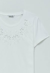 Camiseta de algodón blanca con escote redondo y mangas cortas, que presenta un recorte floral y detalles de lentejuelas en la zona del pecho.