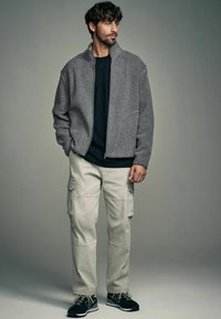 Giacca grigia in pile con zip e superficie testurizzata, abbinata a pantaloni cargo beige e scarpe da ginnastica nere. Design semplice con dettagli sottili.