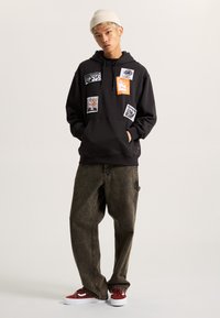 Sudadera negra con parches gráficos en blanco y naranja, combinada con pantalones marrones holgados y zapatillas de ante rojas. Gorro sobre el cabello claro.