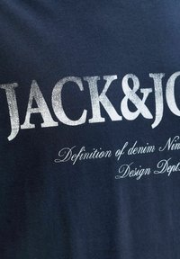T-shirt bleu marine avec un texte blanc audacieux : "JACK&JO", avec un texte plus petit "Definition of denim Nin", sur une texture de tissu lisse.