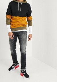 Zwarte pullover hoodie, mosterdgeel en olijfgroen met een voorzak in kangoeroestijl. Draagt bij vervaagde grijze jeans en zwarte sneakers met rode accenten.