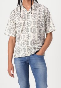 Homme portant une chemise à manches courtes avec des motifs géométriques noirs et un jean bleu, debout avec une main dans la poche sur un fond uni.